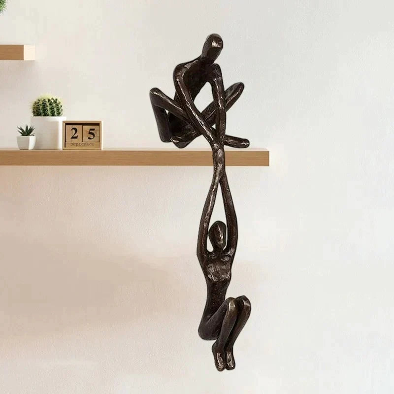 Skulptur i to dele, hvor den ene figur sidder på hyldekanten og holder den anden, bronze eller sort