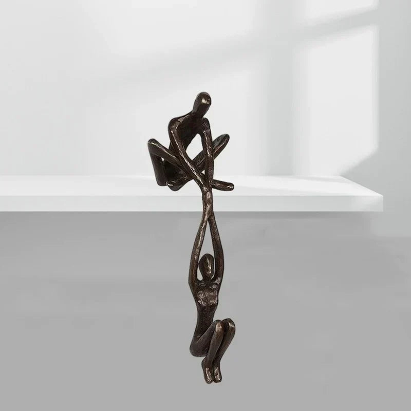 Skulptur i to dele, hvor den ene figur sidder på hyldekanten og holder den anden, bronze eller sort