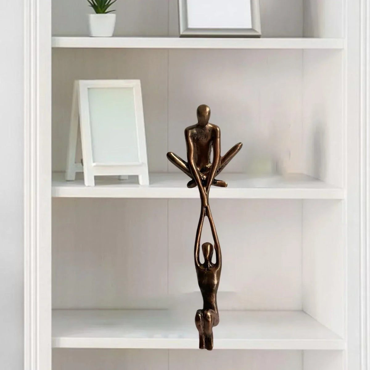 Skulptur i to dele, hvor den ene figur sidder på hyldekanten og holder den anden, bronze eller sort