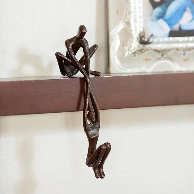Skulptur i to dele, hvor den ene figur sidder på hyldekanten og holder den anden, bronze eller sort