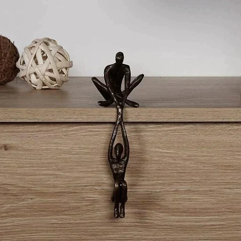 Skulptur i to dele, hvor den ene figur sidder på hyldekanten og holder den anden, bronze eller sort